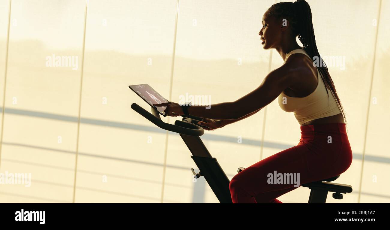 Junge Afrikanerin, die auf einem intelligenten Heimtrainer mit einem Touchscreen-Tablet trainiert. Sie nimmt an einem digitalen Fitnesskurs zu Hause Teil und arbeitet im Schlepptau Stockfoto