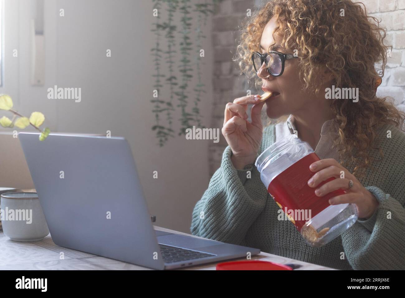 Eine Frau, die einen Laptop auf dem Tisch benutzt und Kekse aus der Box isst. Menschen, die Müll essen und gleichzeitig arbeiten. Beschäftigte Person, die während des Frühstücks oder der Pause am Computer arbeitet. Moderner Lebensstil Stockfoto