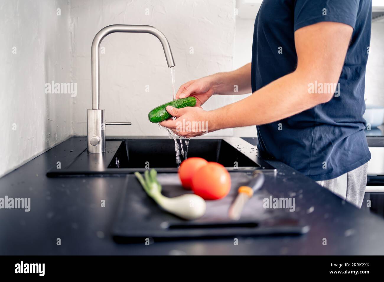 Gemüse im Spülbecken mit Wasser waschen. Lebensmittelzubereitung. Mann kocht. Salatzubereitung. Gurke in der Hand unter Wasserhahn. Vegan oder vegetarisch. Stockfoto