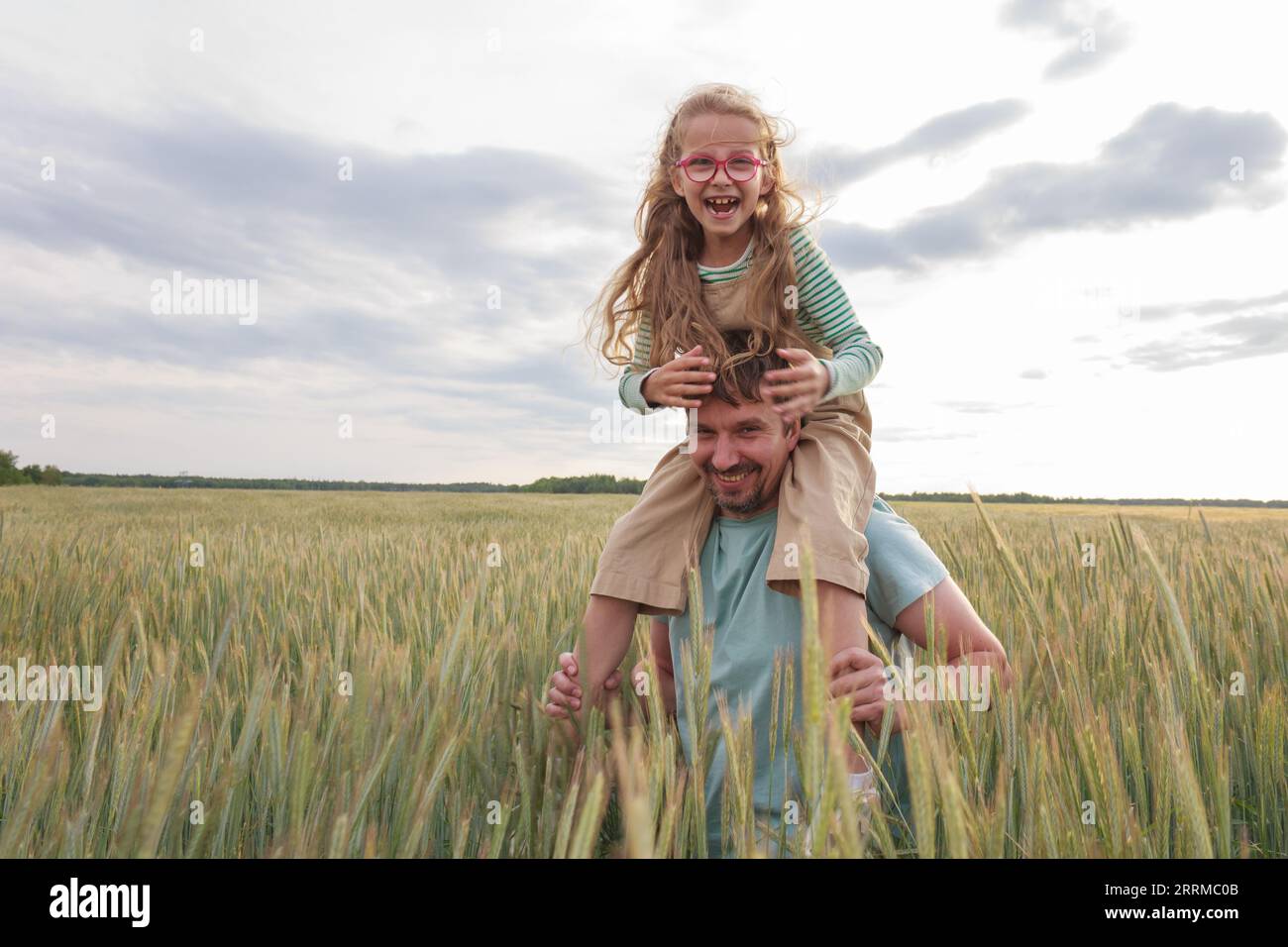 Eine Tochter, die auf den Schultern ihres Vaters vor dem Hintergrund eines Weizenfeldes sitzt. Glückliche Momente in der Natur. Stockfoto