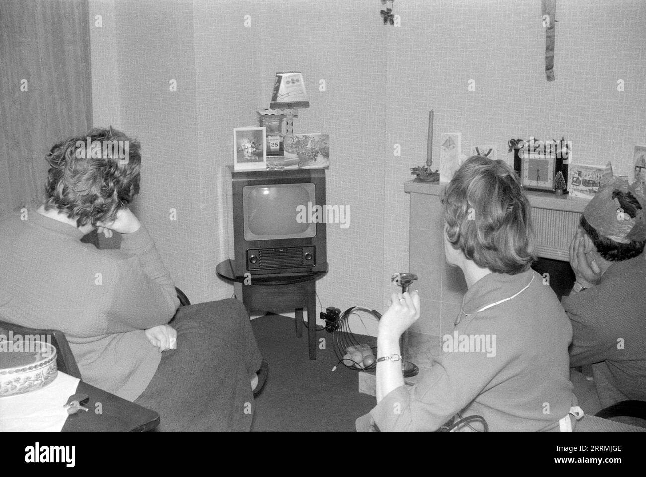 England. c.1960 – Eine Familie sitzt am ersten Weihnachtsfeiertag in einer Lounge und schaut Fernsehen. Der Fernseher mit Holzgehäuse ist ein TV43-Modell, das von Bush in den 1950er Jahren hergestellt wurde Der Mann trägt einen Papierhut von einem Weihnachtskracker. Auf dem Fernseher befindet sich ein Kalender, der das Datum des 25. Dezember anzeigt. Das Zimmer hat einen gefliesten Kamin umgeben, der mit Grußkarten, Kerzen und holly dekoriert ist. Stockfoto