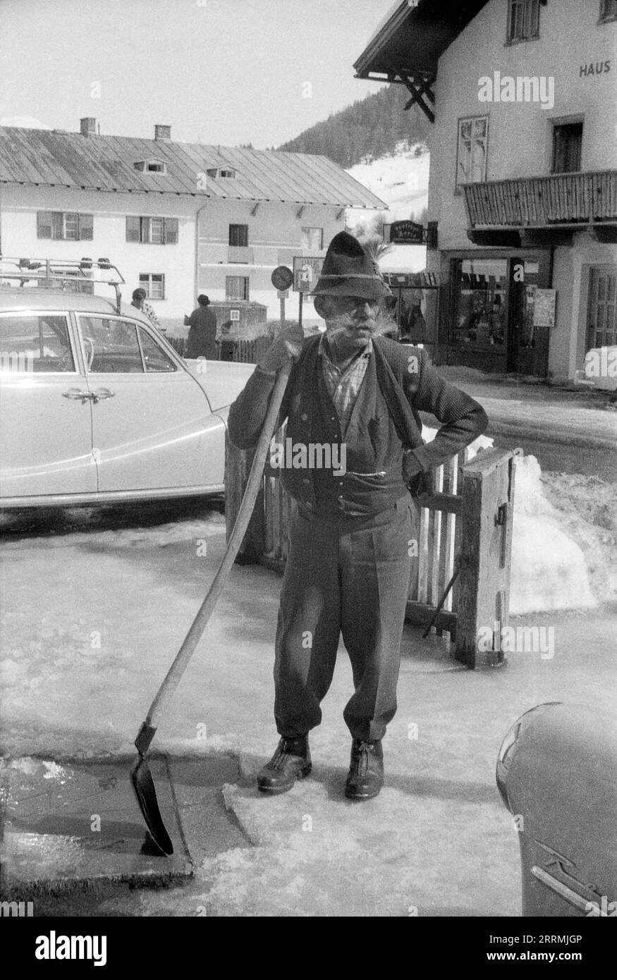 Tirol, Österreich. c.1960 – St. Anton am Arlberg. Ein älterer Mann mit einem buschigen Hammelbart und einem traditionellen Tiroler Hut steht mit einer langgezogenen Schaufel, mit der er Schnee vom Pflaster entfernt. Hotels und die schneebedeckten Berge der Ostalpen sind in der Ferne sichtbar. Stockfoto