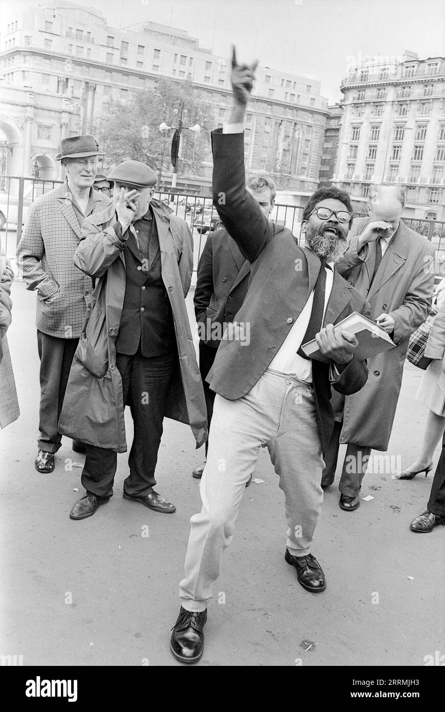 London. 1965: Ein Evangelist predigt in Speaker’s Corner, Hyde Park an der Kreuzung von Cumberland Gate, in der Nähe von Marble Arch, London. Der Prediger hält eine Bibel in der einen Hand und zeigt seine andere Hand zum Himmel. Eine Gruppe von Zuschauern hat sich um ihn herum versammelt. Ein älterer Mann, der einen Regenmantel und eine flache Kappe trägt, verspottet den Prediger, indem er ihm eine Himbeere durch die Hand bläst. Das Cumberland Hotel und das Gebäude an der 140 Park Lane sind in der Ferne zu sehen. Stockfoto