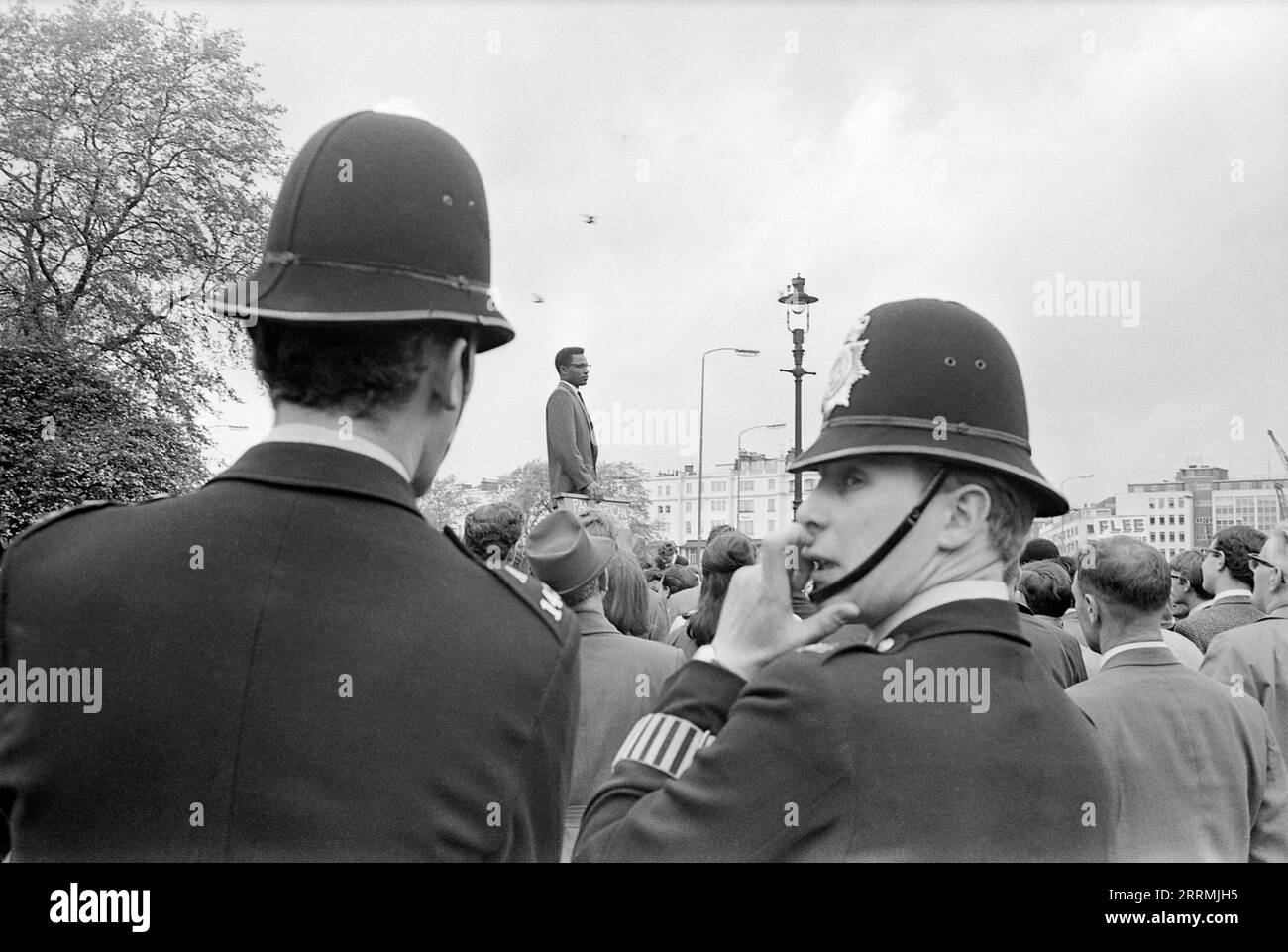London. 1965: Ein Mann, der auf einer Plattform steht und den Panafrikanischen Nationalkreuzzug vertritt, spricht in der Speaker’s Corner im Hyde Park, London, vor einer Menge von Zuschauern. Zwei Metropoliten stehen im Vordergrund, einer schaut über seine Schulter, das Pflichtarmband auf seinem Ärmel ist zu sehen. Stockfoto