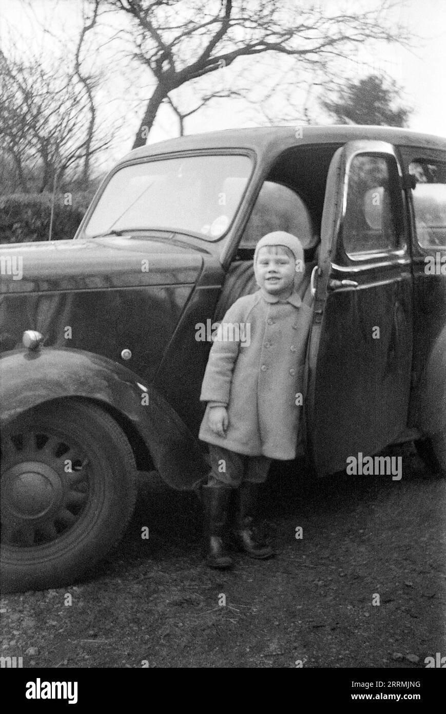 England. c.1960: Ein kleiner Junge mit Mantel, gummistiefeln und Wollmütze steht vor der offenen Tür eines Standard-Acht-Limousine-Wagens aus dem Jahr 1948. Stockfoto