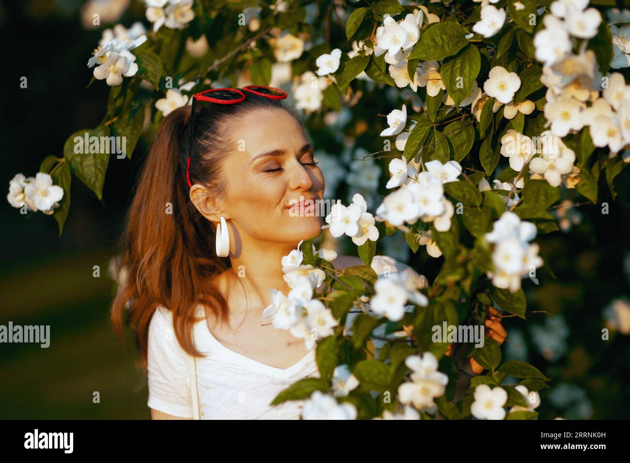 Sommerzeit. Glückliche moderne 40-jährige Frau in weißem Hemd in der Nähe von blühendem Baum. Stockfoto