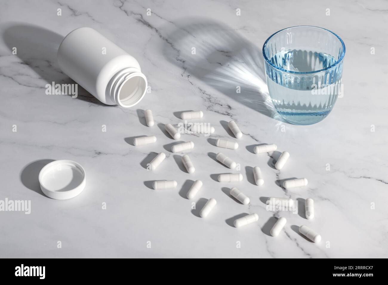 Weiße medizinische Pillen und ein Glas Wasser auf einem grauen Marmortisch Hintergrund. Stillleben mit harten Schatten. Ein Haufen verstreuter Kapseln. Gesundheitsfürsorge konc Stockfoto