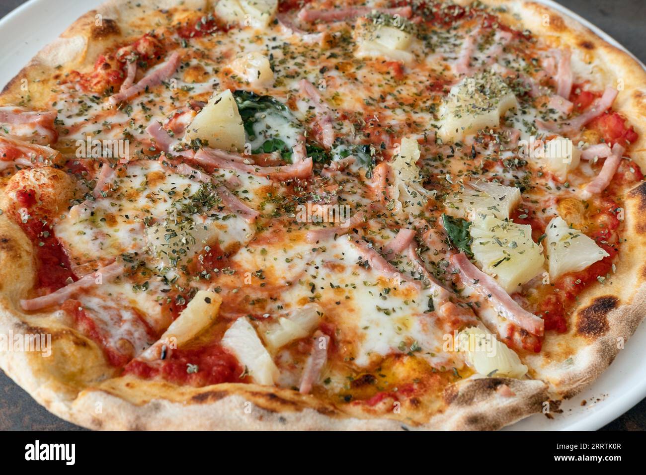 Nahaufnahme einer ganzen Pizza aus Ananas und Schinken mit dünner Kruste auf einem Teller voller köstlicher Zutaten und mit aromatischem getrocknetem Oregano bestreut Stockfoto