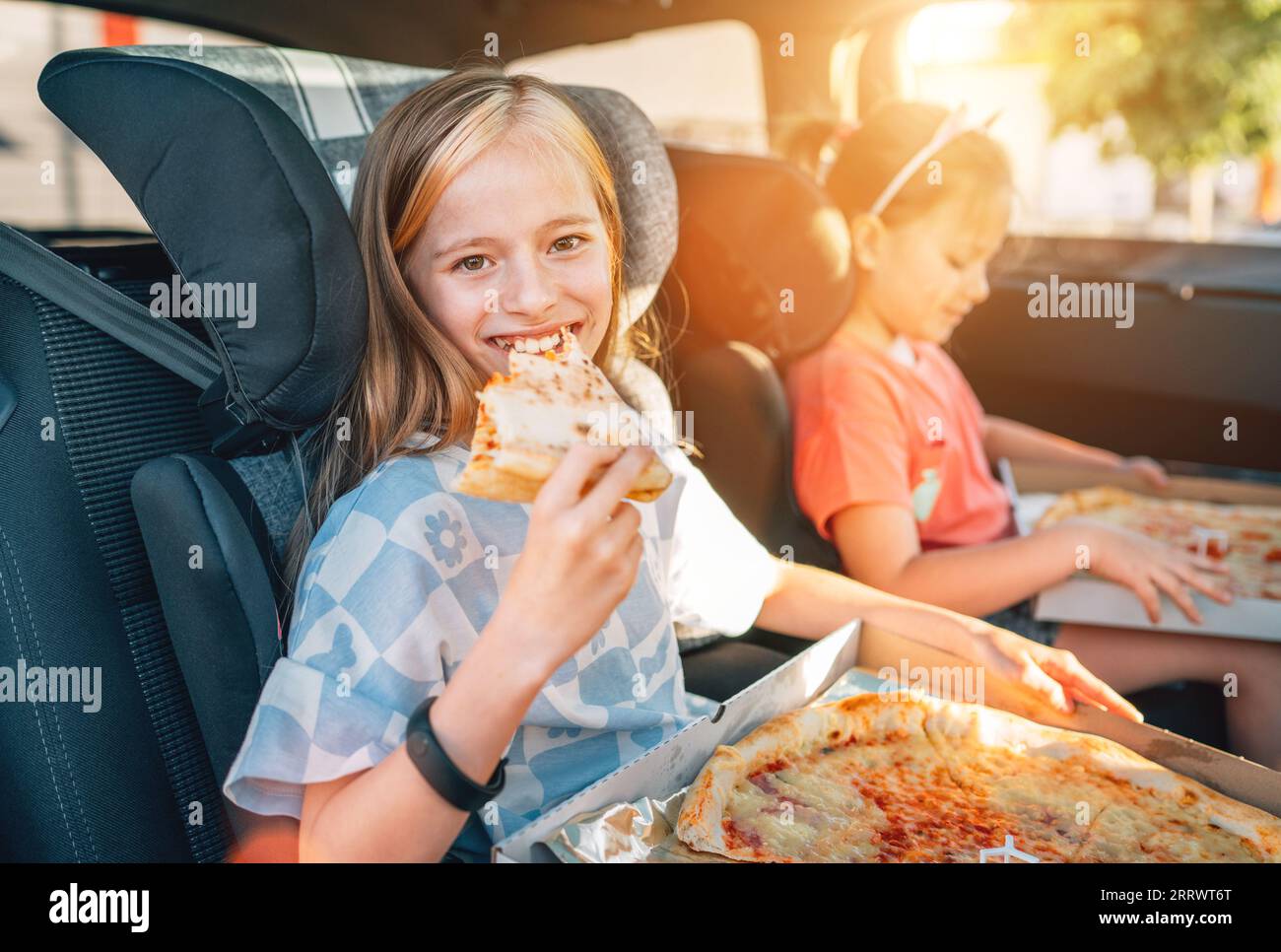 Portrait eines lächelnden Mädchens, das gerade gekochte italienische Pizza isst, sitzt mit einer Schwester auf dem Rücksitz in Kindersitzen. Fröhliche Kindheit, Fastfoo Stockfoto
