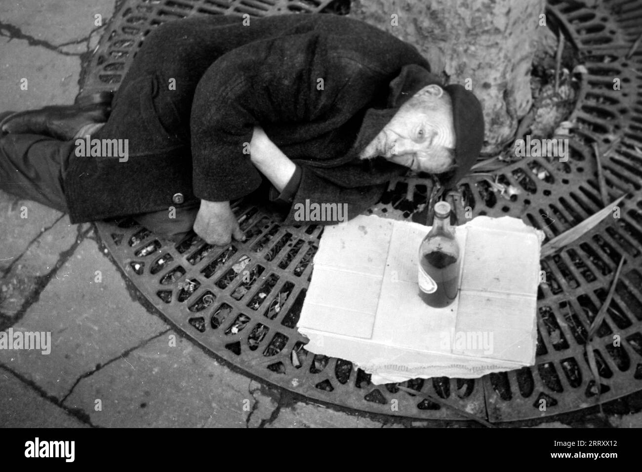 Ein obdachloser Mann liegt an einen Baum gelehnt auf einem Pariser Trottoir, 1962. Ein Obdachloser liegt auf einem Pariser Pflaster, 1962, an einem Baum. Stockfoto