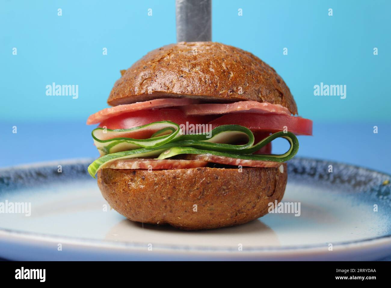 Ein Burger-Sandwich mit Wurstgemüse Gurkentomaten mit Buchweizen-Roggenbrötchen liegt auf einem blauen Teller auf blauem Hintergrund. Vertikales Foto. Kirsch Stockfoto