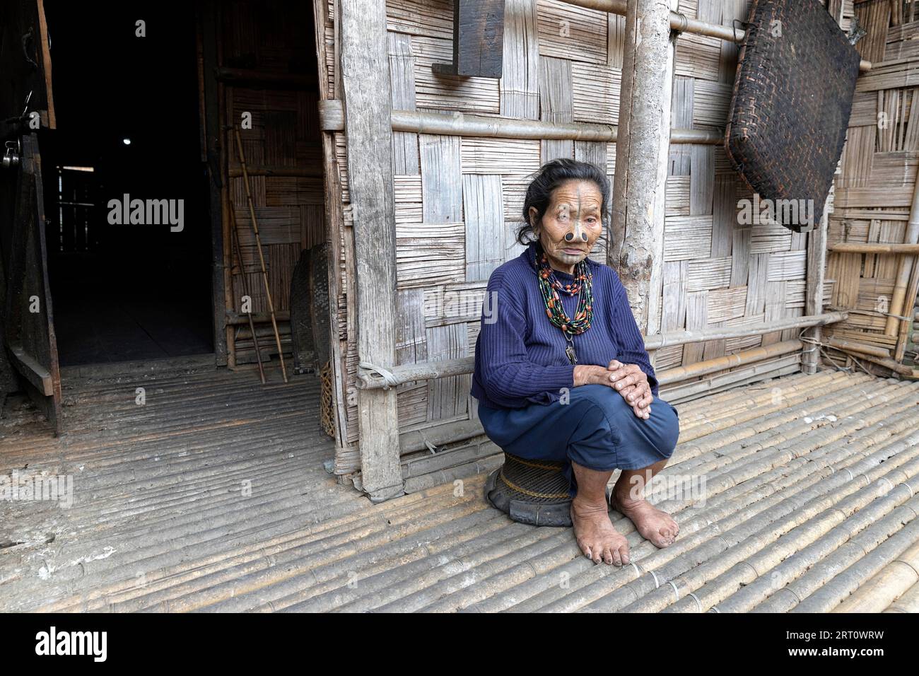 Die einheimische Frau aus dem Stamm der Apatani sitzt auf einem kleinen Stuhl vor einem traditionellen Holzhaus in einem kleinen Dorf in der Nähe von Ziro in Arunachal Pradesh, Indien Stockfoto