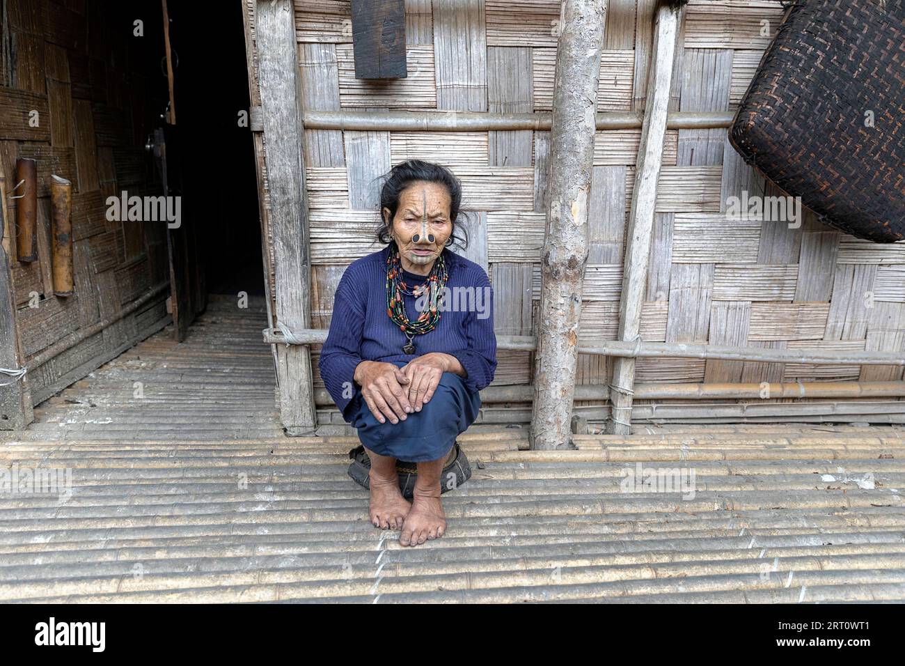 Die einheimische Frau aus dem Stamm der Apatani sitzt auf einem kleinen Stuhl vor einem traditionellen Holzhaus in einem kleinen Dorf in der Nähe von Ziro in Arunachal Pradesh, Indien Stockfoto