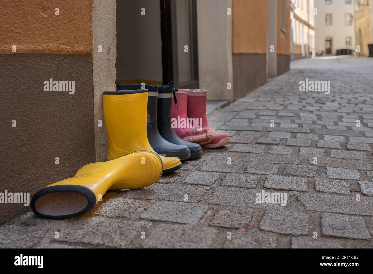 Drei Paar Gummistiefel für Kinder in verschiedenen Farben stehen auf der Straße neben der Haustür in einer städtischen Gasse mit Kopfsteinpflaster Stockfoto