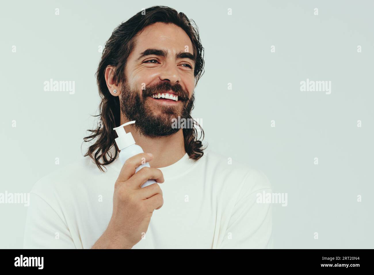 Mann mit einem Bart, der glücklich mit seiner Wahl in einem Hautreiniger aussieht, lächelt, während er in einem Studio wegschaut. Gutaussehender junger Mann mit gesundem Anti-Aging Stockfoto