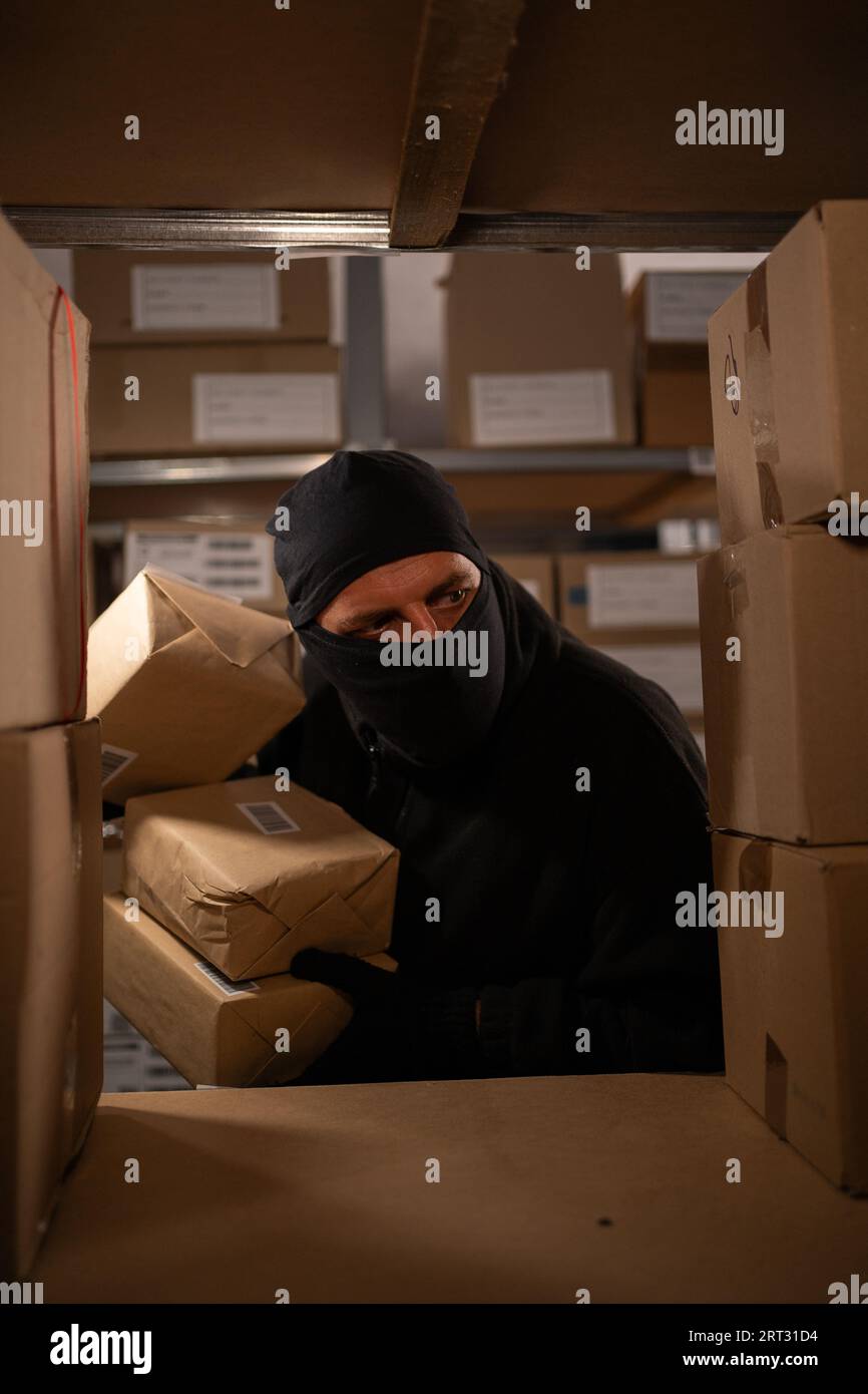 Ein nächtlicher Dieb in einer schwarzen Maske stiehlt in einem Lagerhaus Kisten mit Waren. Zur Problematik des Diebstahls von Gütern Stockfoto