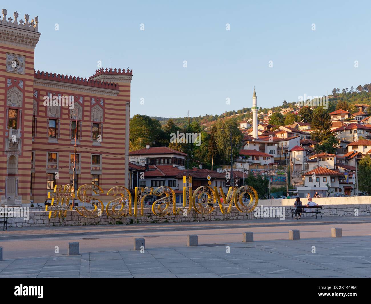 Die Menschen sitzen auf einer Bank neben dem Sarajevo-Schild mit dem Rathaus links und den Häusern auf einem Hügel in Sarajevo, Bosnien und Herzegowina, 10. September 2023 Stockfoto