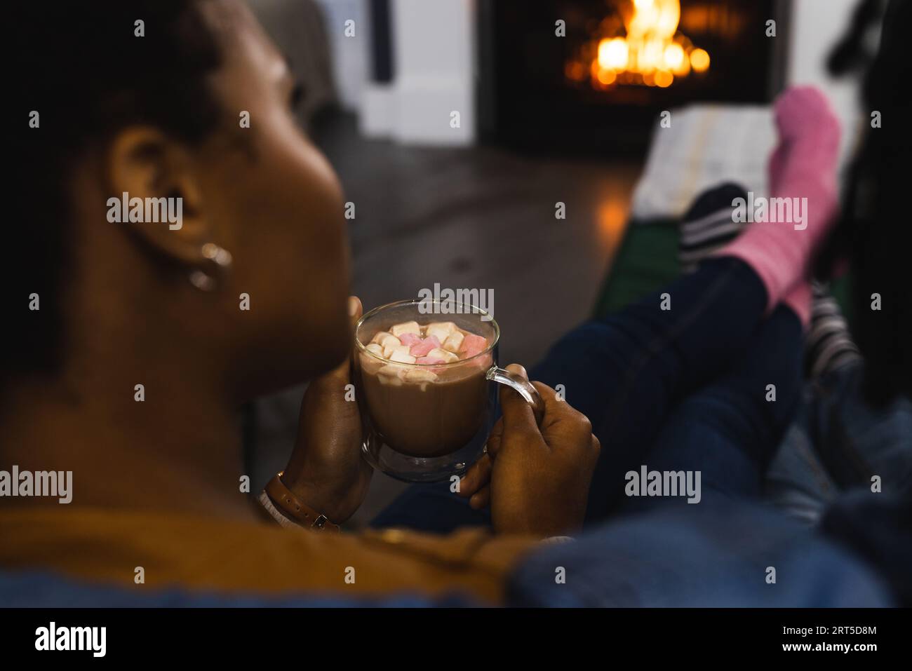 afroamerikanisches Paar in Socken, das sich zu Hause vor dem offenen Feuer mit heißer Schokolade entspannt Stockfoto