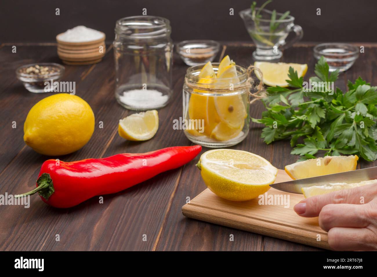 Hacken Sie mit den Händen eine Zitrone auf dem Schneidebrett. Zitrone im Glas. Chilischote, ganze Zitrone und Salz. Fermentierte Lebensmittel zur Stärkung der Immunität. Dunkler Holzhintergrund Stockfoto