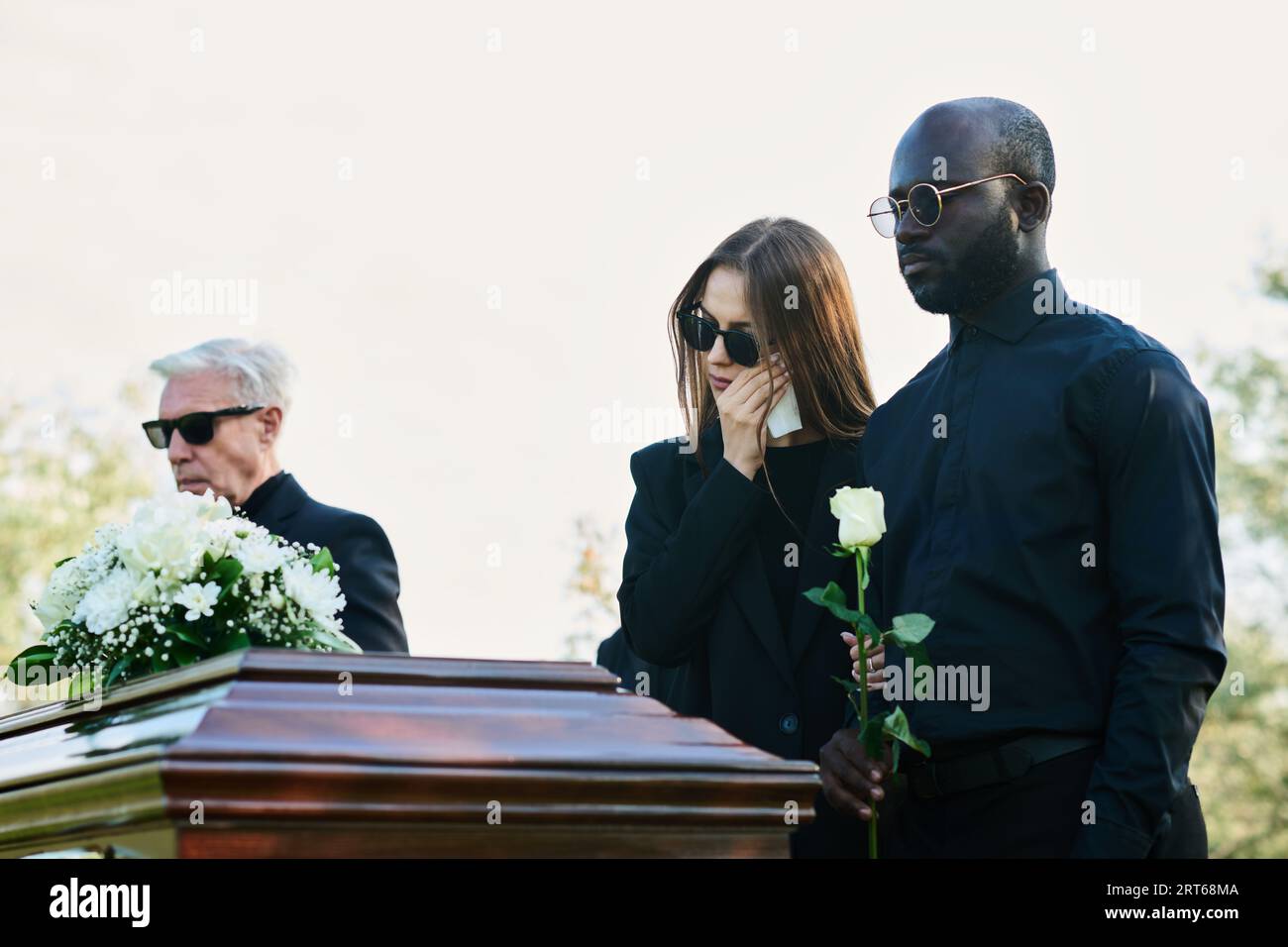 Junger Trauermann mit weißer Rose, die neben seiner Frau steht, in  schwarzer Kleidung und Sonnenbrille, die bei der Beerdigung des  Familienmitglieds Tränen mit einem Taschentuch abwischt Stockfotografie -  Alamy