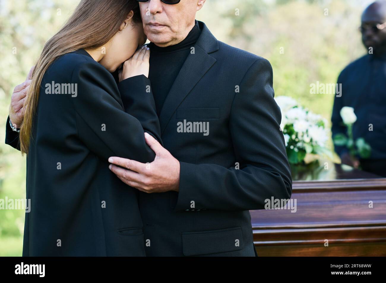 Reifer trauernder Vater umarmt die trauernde Tochter, die auf seiner Schulter weint, während beide bei der Beerdigung gegen Sarg mit geschlossenem Deckel stehen Stockfoto