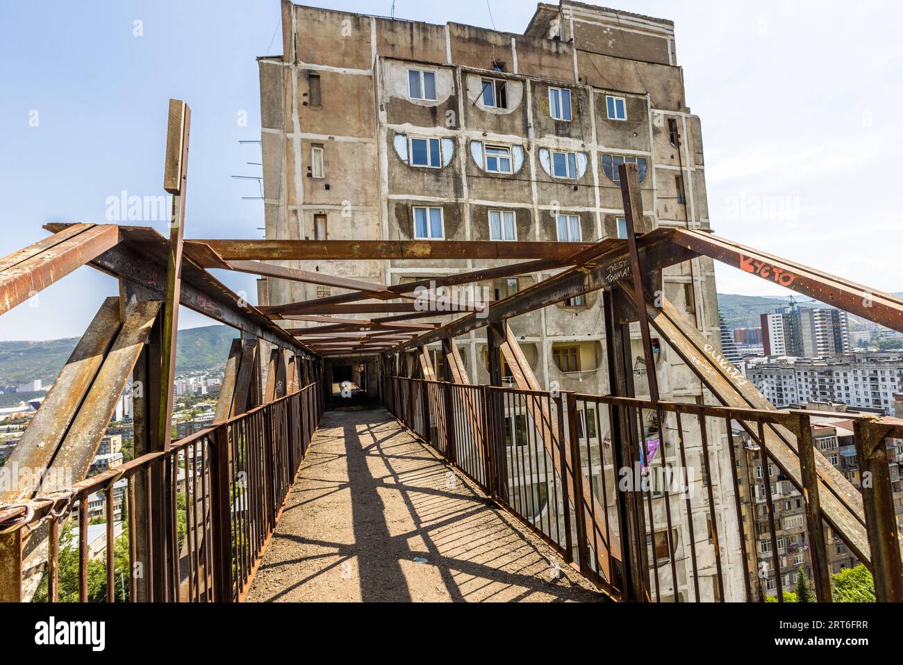 Ein Gehweg des Wohnkomplexes Tbilisi Skybridge (Nutsubidze Skybridge) in der Ermile-Bedia-Straße in Tbilissi, Georgien, führt zwischen rostigen Stahlträgern zu einem freiliegenden Betonturmblock. Seitenzäune schützen den Gehweg vor dem Wind. Weiß gerahmte, hufeisenförmige Fensteröffnungen strukturieren die Fassade. Rechts liegen Stadthochhäuser, links bewaldete Hügel. Der Komplex verbindet die Stadtteile Saburtalo und Nutsubidze am Hang. Er liegt etwa 11 Kilometer vom historischen Stadtzentrum entfernt. Die Architekten Otar Kalandarishvili und Gaioz Potskhishvili entwarfen ihn 1974 als den Sha Stockfoto