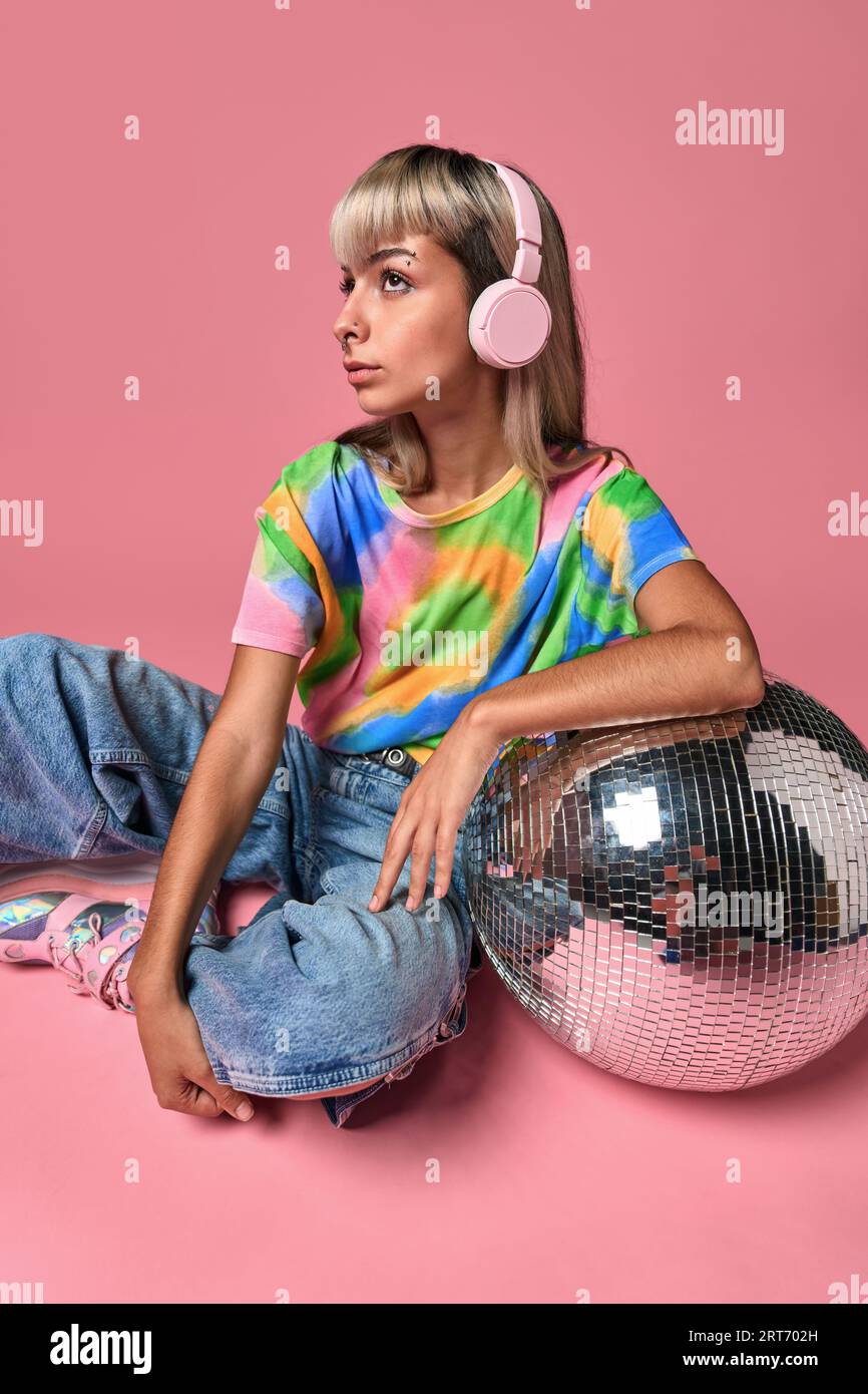 Junger Hipster im Street-Style, der Kopfhörer berührt, während er Musik auf einem Disco-Ball vor rosafarbenem Hintergrund hört Stockfoto