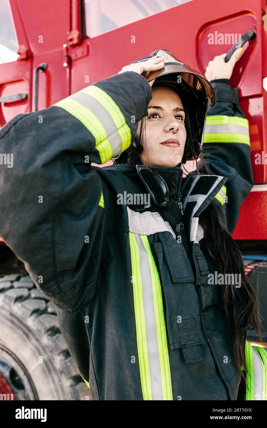 Junge, furchtlose Feuerwehrfrau trägt Arbeitskleidung, die den Schutzhelm anpasst, während sie sich auf die Arbeit vorbereitet, um in die rote Feuerwehrmaschine zu gelangen Stockfoto