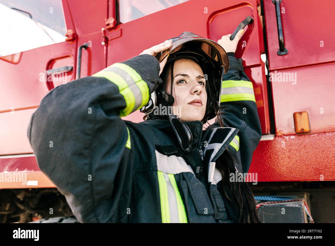 Junge, furchtlose Feuerwehrfrau trägt Arbeitskleidung, die den Schutzhelm anpasst, während sie sich auf die Arbeit vorbereitet, um in die rote Feuerwehrmaschine zu gelangen Stockfoto