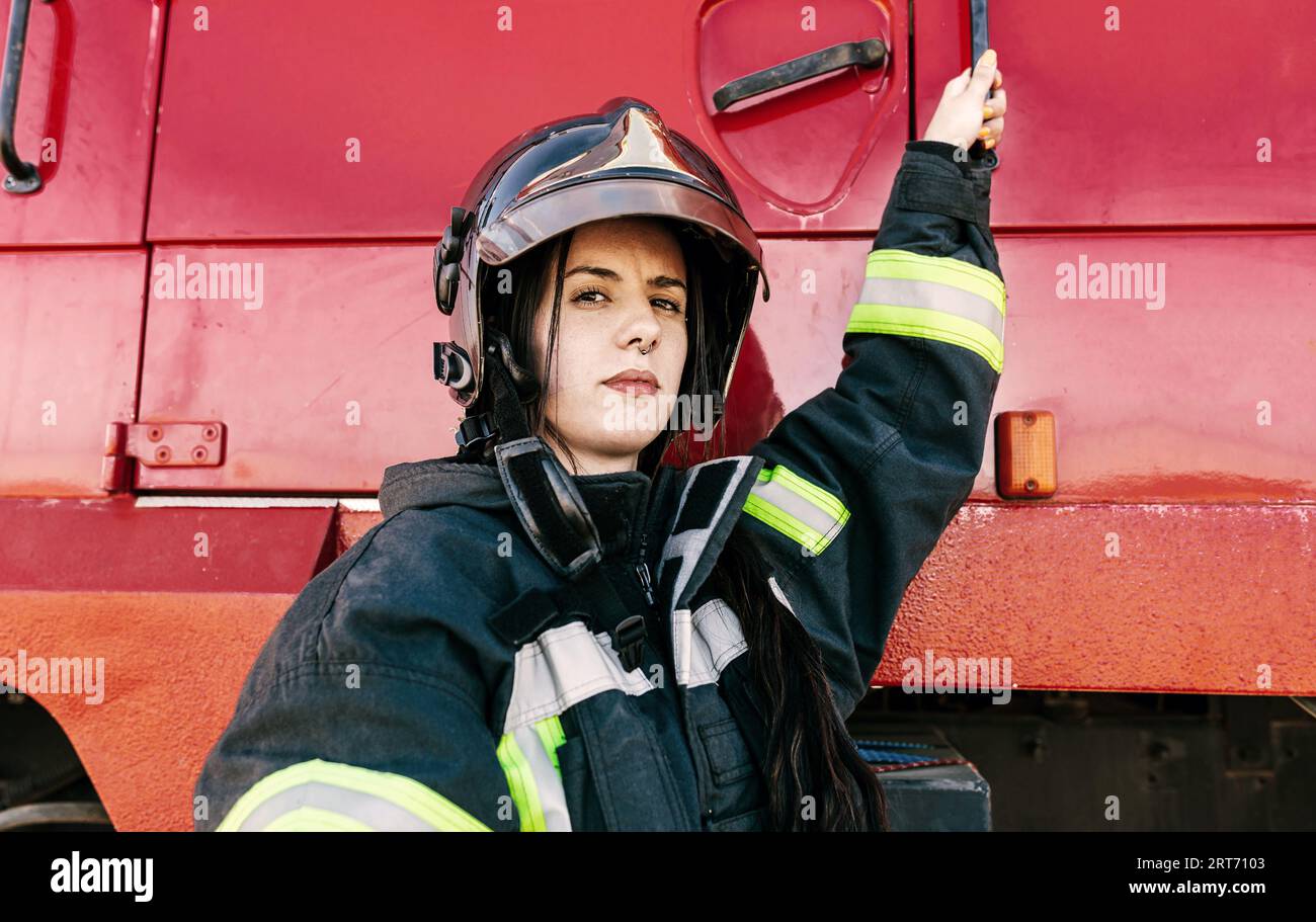 Junge furchtlose Feuerwehrfrau, die Arbeitskleidung und Schutzhelm trägt, um sich auf die Arbeit vorzubereiten und in die rote Feuerwehrmaschine zu gelangen Stockfoto
