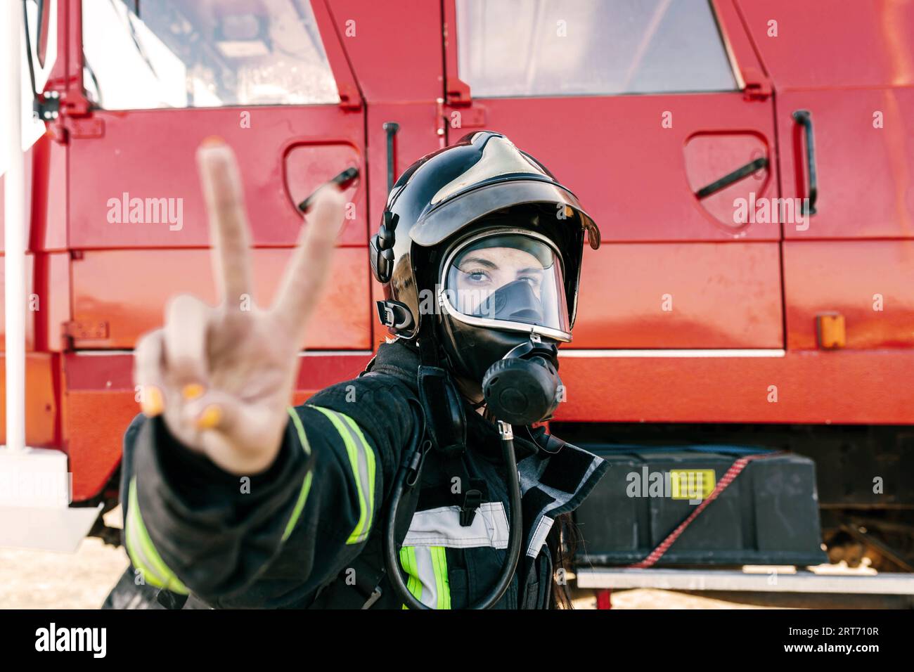 Feuerwehrfrau in Schutzuniform und Atemschutzmaske mit Siegeszeichen, während sie in der Nähe des roten Lkws steht und die Kamera anschaut Stockfoto