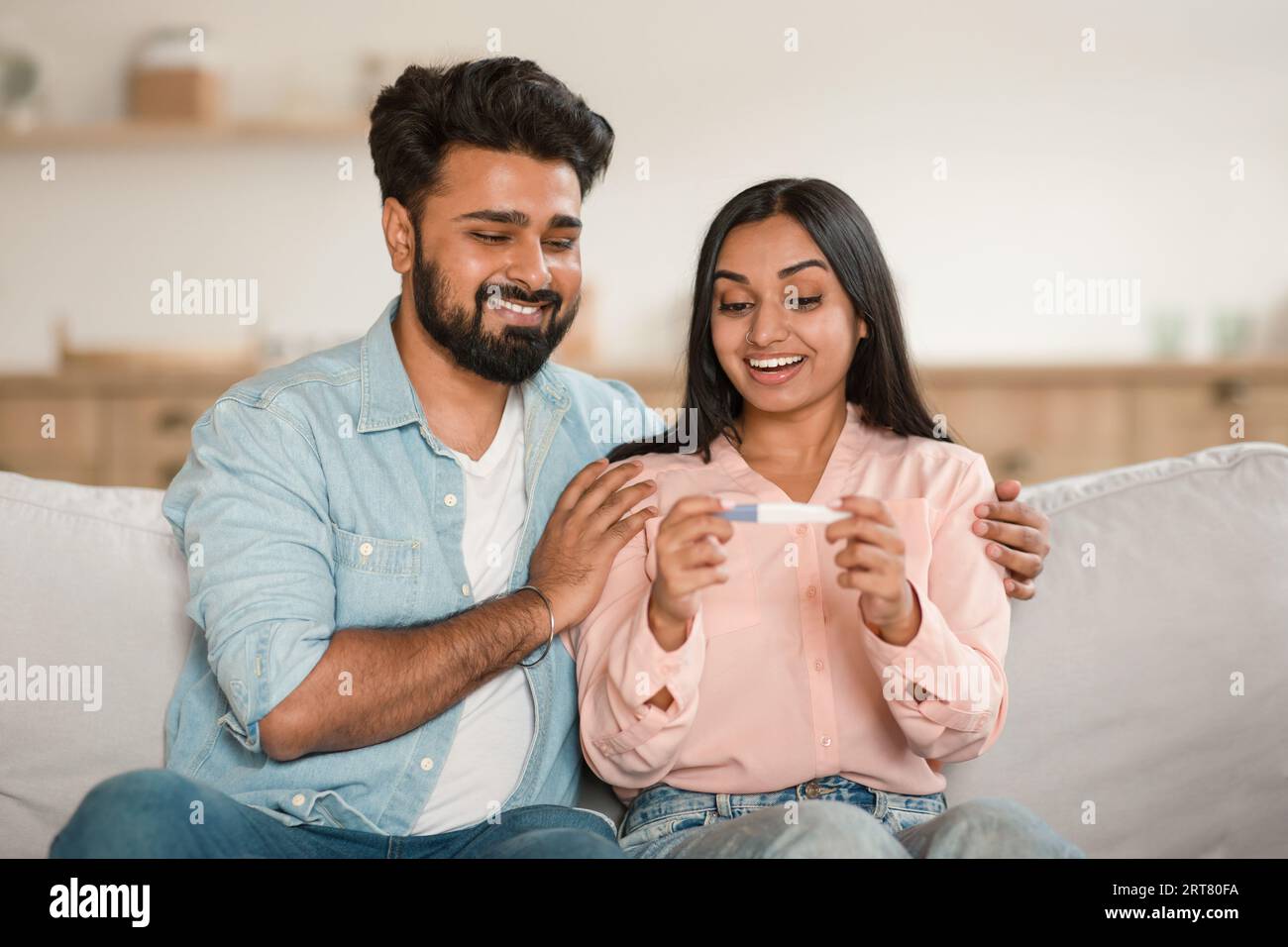 Glückliche junge indische Ehepartner, die sich einen positiven Schwangerschaftstest ansehen, aufgeregt, Eltern zu werden, zu Hause auf der Couch sitzen Stockfoto