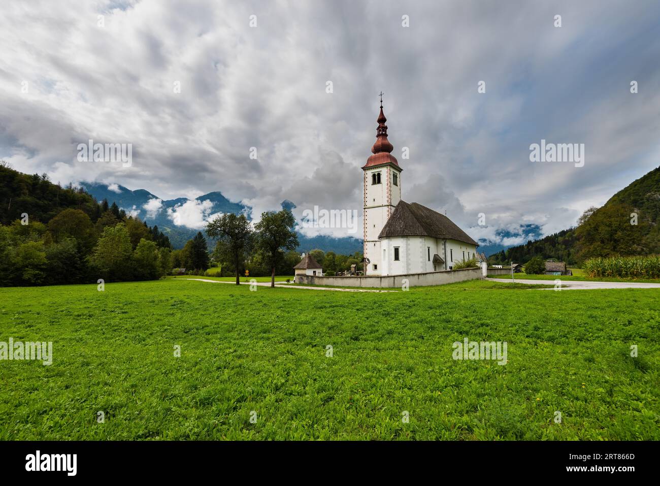 Schöne alte einsame Kirche mit angrenzendem Friedhof im Bergtal Mit frischem Gras und bewölktem Himmel inmitten hoher Berggipfel Stockfoto