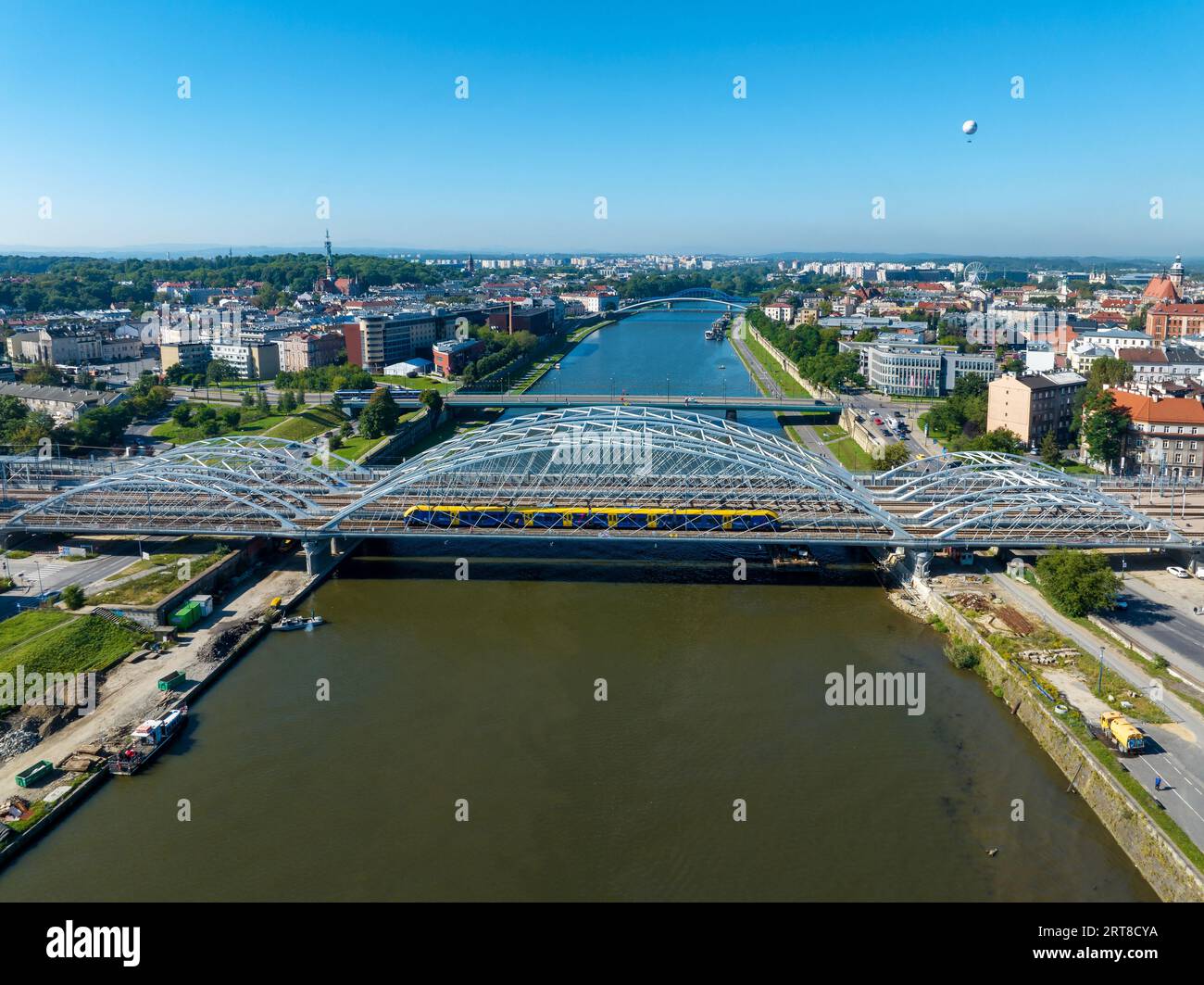 Neue dreifach gebundene Eisenbahnbrücke mit laufendem Zug über die Weichsel in Krakau, Polen, weitere Brücken, Altstadt und touristischer Beobachtungsballon Stockfoto