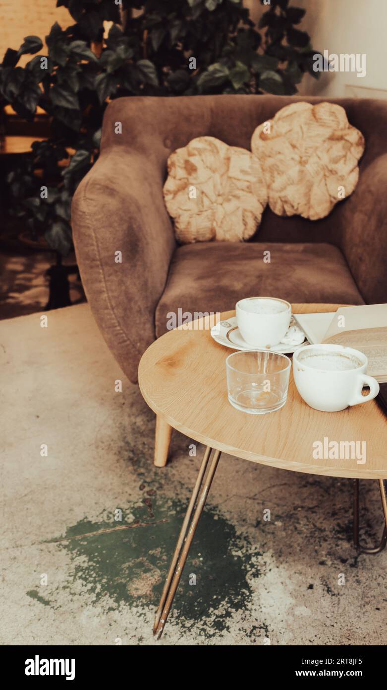 Kaffeetassen und Buch auf Holztisch. Sessel mit Kissen und Tisch mit Kaffeetassen im Café. Frühstückstisch im Restaurant. Moderner urbaner Lebensstil Stockfoto