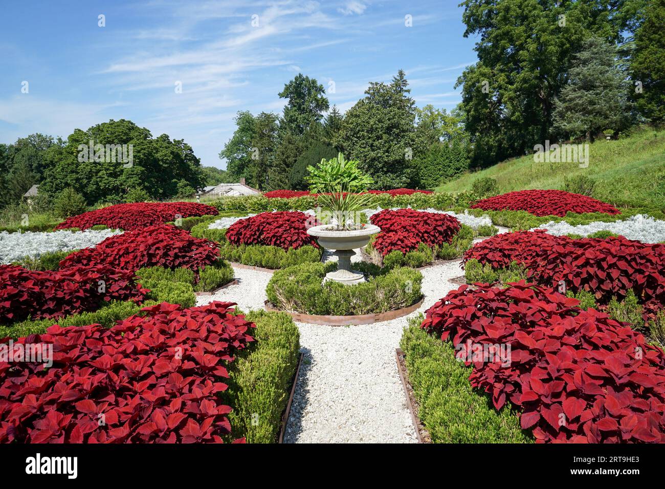 Wunderschöner Blick auf einen formellen Garten mit einer großen Steinurne voller Pflanzen. Es gibt einen weißen Steinsteg, der von üppigem Grün umgeben ist und tief ist Stockfoto