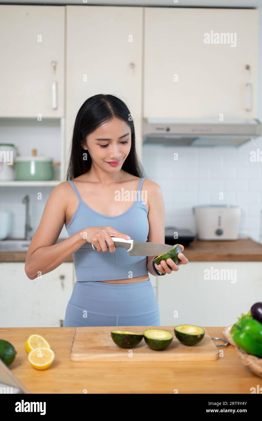 Eine schöne und gesunde asiatische Frau in Turnkleidung schneidet eine Avocado und bereitet ihr gesundes Frühstück in der Küche vor, bevor sie ins Fitnessstudio geht. Heilen Stockfoto