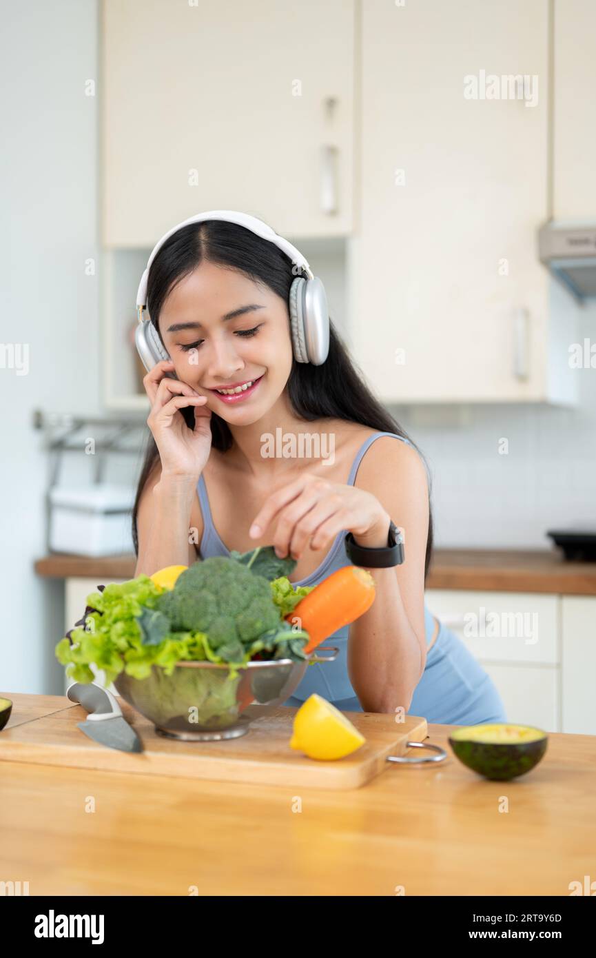 Ein Porträt einer hübschen asiatischen Frau in Turnkleidung genießt die Musik auf ihren Kopfhörern, während sie Zutaten für ihr gesundes Frühstück und Smoothi zubereitet Stockfoto