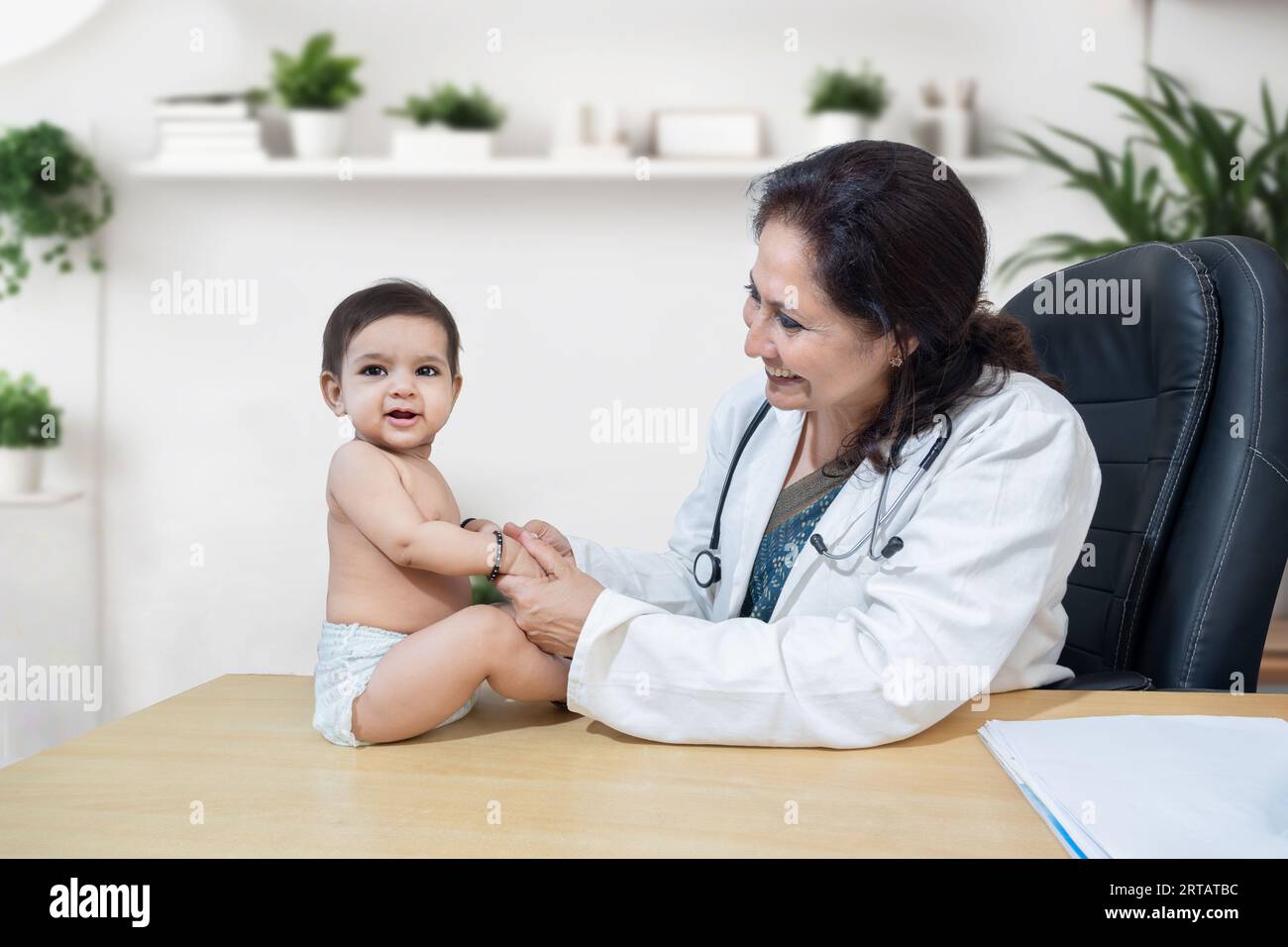 Junge lächelnde indische Ärztin, die in ihrer Klinik einen Check-up für kleine Kleinkinder macht. Regelmäßige Untersuchungen zum Kinderarzt. Kinderheilkunde und Stockfoto
