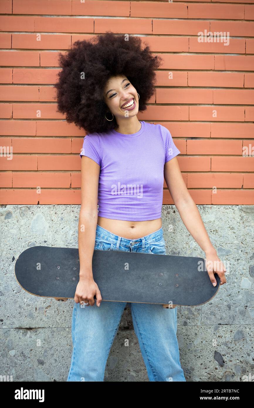 Positive afroamerikanische Frau mit Afro-Frisur und in lässiger Kleidung, die Kamera anschaut, während sie sich auf eine Ziegelwand mit Skateboard lehnt Stockfoto