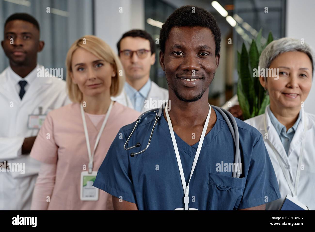 Junge lächelnde afroamerikanische männliche Assistentin in blauer Uniform mit Blick auf die Kamera, während sie gegen eine Gruppe interkultureller Kollegen steht Stockfoto