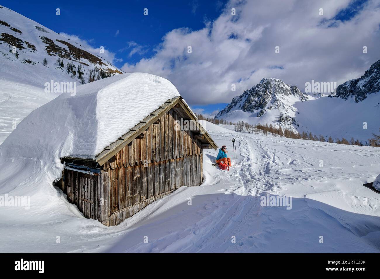 Frau auf einer Skitour sitzt auf einer schneebedeckten Alm mit Bielschitza im Hintergrund, Matschacher Alm, Rosental, Karawanken, Kärnten, Österreich Stockfoto