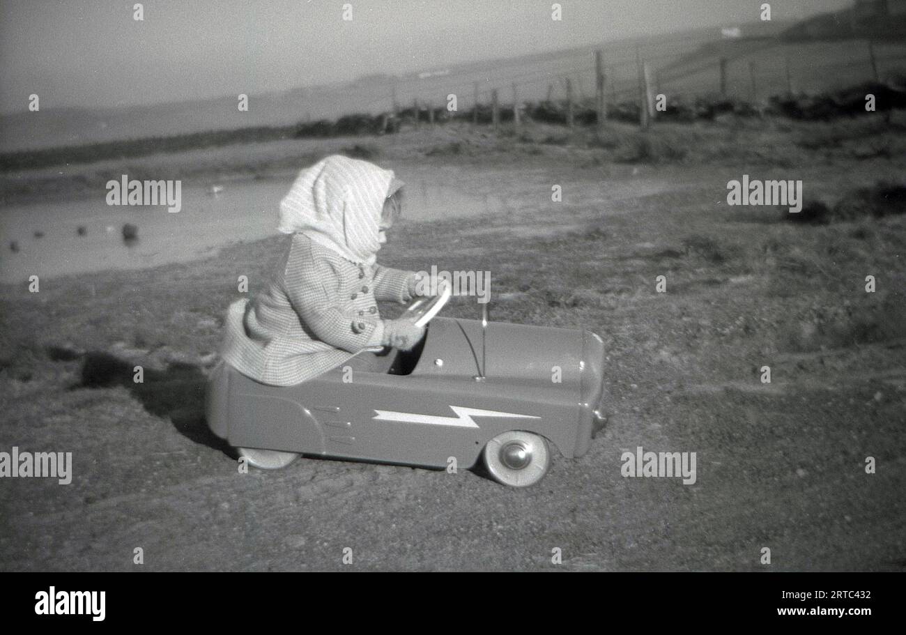 1950er Jahre, historisch, draußen auf offenem Gelände, in einem grasbewachsenen Gebiet an einem Naturteich, ein kleines Mädchen mit Mantel und Kopftuch, das in einem Spielzeugwagen fährt/spielt, England, Großbritannien. Stockfoto