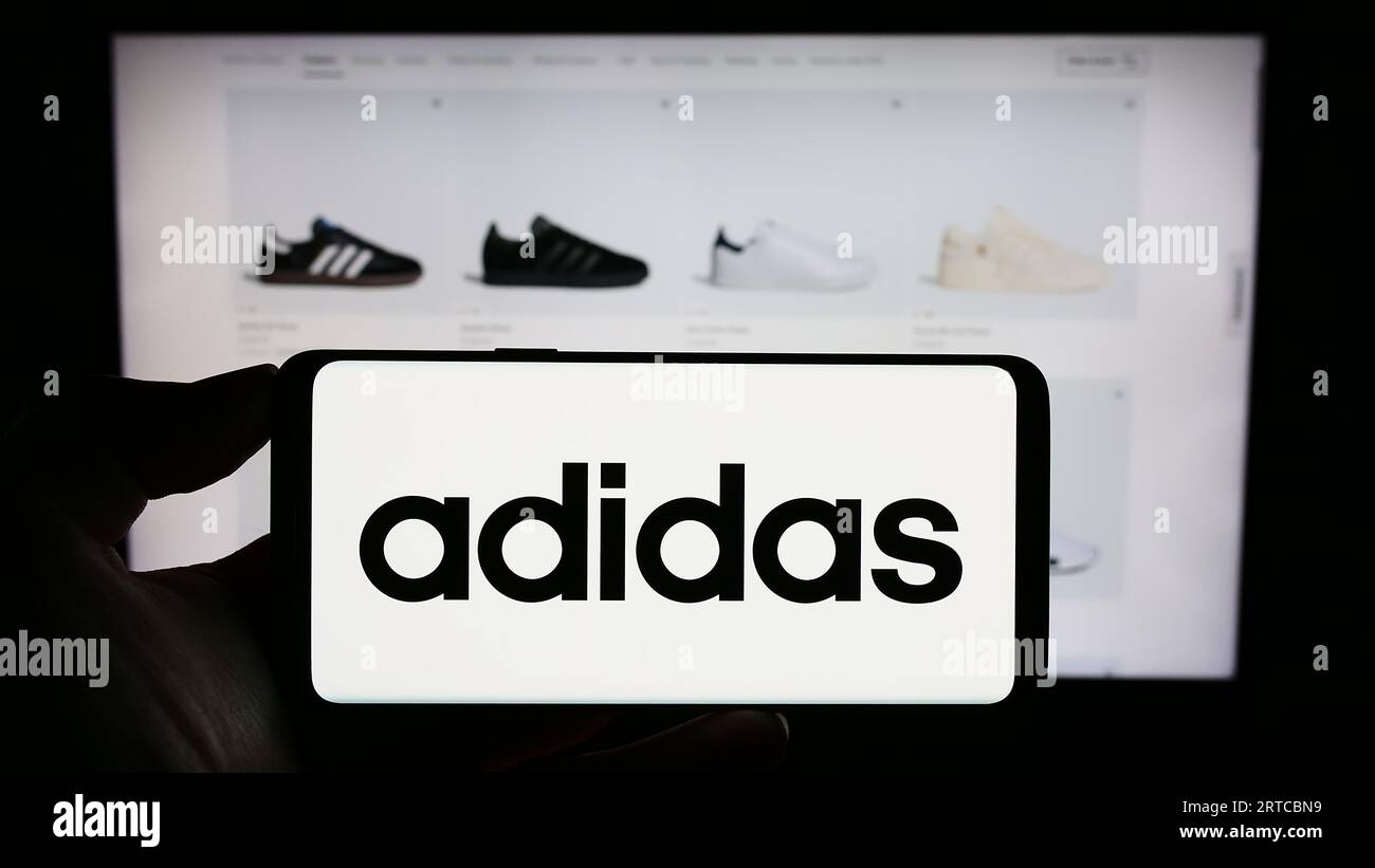 Person, die ein Mobiltelefon mit dem Logo des deutschen Bekleidungs- und Schuhherstellers Adidas AG auf dem Bildschirm vor der Webseite hält. Konzentrieren Sie sich auf das Display des Telefons. Stockfoto