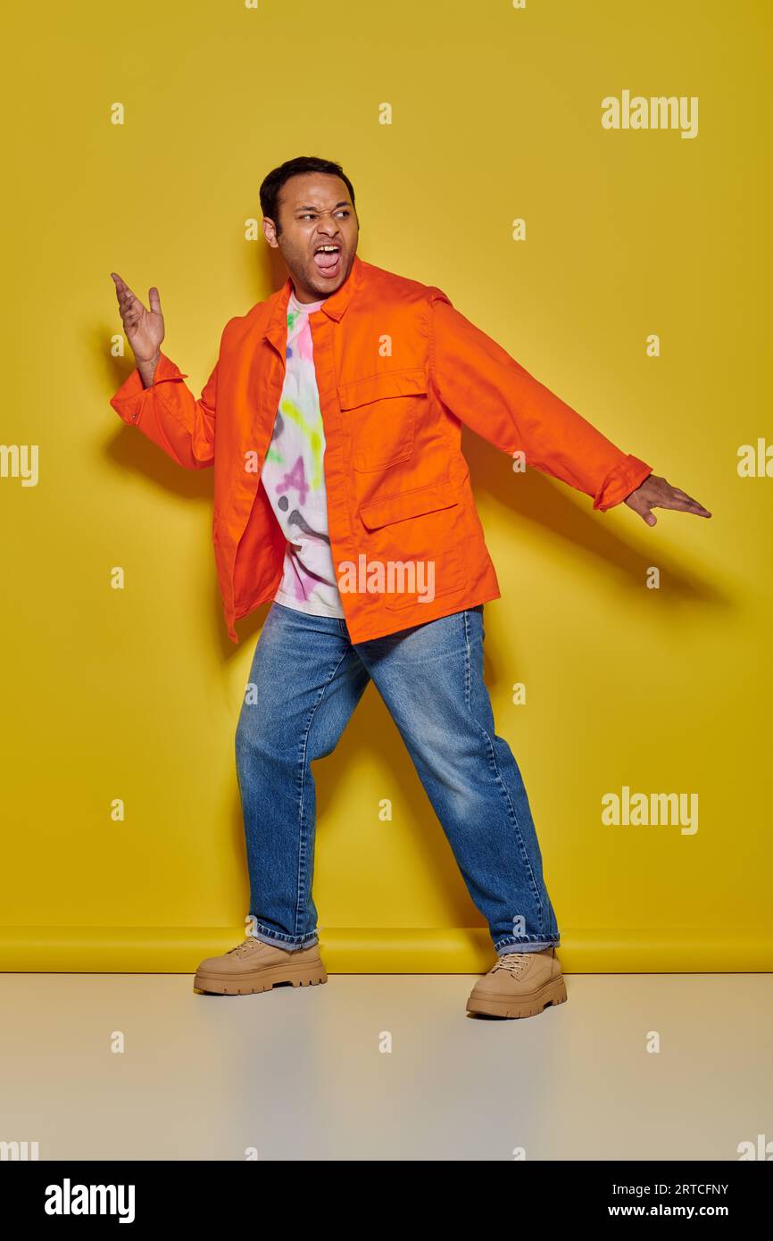Emotionaler indischer Mann in orangefarbener Jacke und Jeans, der vor gelbem Hintergrund gestikuliert und schreit Stockfoto