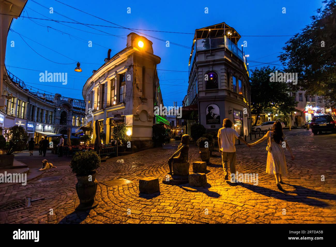 Eine kleine Bronzestatue, möglicherweise die Tamada-Statue, steht auf einem runden Block in der Sioni-Straße in der Altstadt von Tiflis, Georgien, am Abend. Der Tamada ist der Toastmeister, der über das georgische Festmahl namens Supra vorsitzt. Zwei kleinere Blöcke stehen neben der Figur. Ein Paar geht Hand in Hand über die beleuchteten Kopfsteinpflastersteine. Der Mann trägt ein weißes T-Shirt, die Frau ein langes helles Kleid. Links liegt ein Hund vor einem Eckgebäude mit hell erleuchteten Fenstern. Ein großes Porträt eines Mannes mit Hut hängt an der gegenüberliegenden Fassade. Straßenlaternen und Kabel erstrecken sich unter dem tiefen Stockfoto