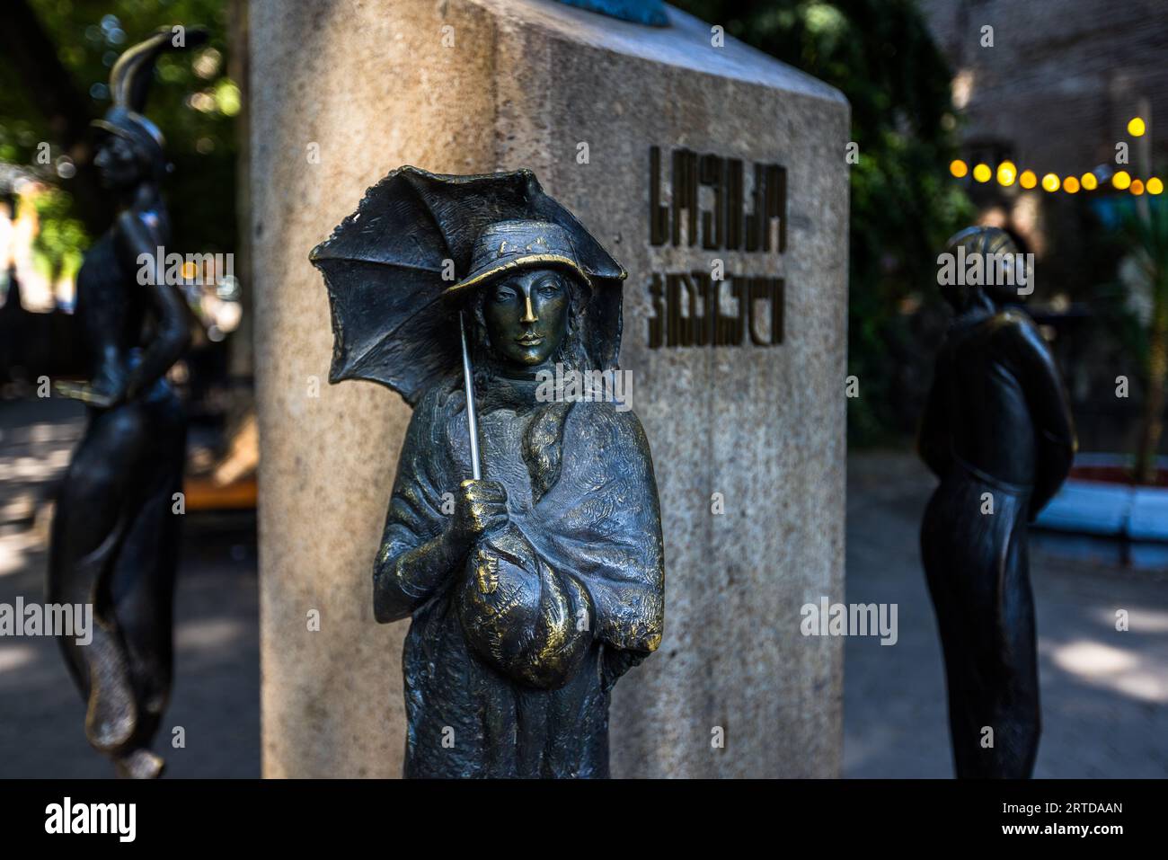 Eine Bronzestatue einer Frau mit Hut und offenem Regenschirm ist Teil des Denkmals für die Schauspielerin Sofiko Chiaureli im Sofiko-Chiaureli-Garten in der Kote-Abkhazi-Straße in der Altstadt von Tbilissi, Georgien. Die Figur trägt einen Schal um die Schultern und hält eine Tasche vor sich. Dahinter steht ein steinerner Sockel mit georgischer Schrift, flankiert von Bronzestatuen von Frauen links und rechts. Das Denkmal des Bildhauers Levan Vardosanidze wurde 2009 enthüllt. Vier Figuren umgeben einen Büsten und zeigen die Schauspielerin in verschiedenen Rollen. Sofiko Chiaureli (1937–2008) spielte in mehr als 100 Stockfoto