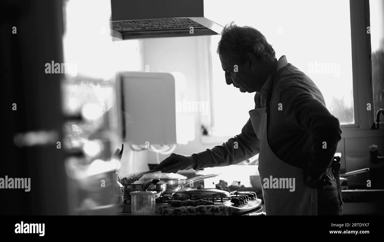 Einfarbige Szene, in der älterer Koch das Essen auf dem Küchenherd umrührt, offener älterer Mann, der sich auf die Zubereitung von Speisen von Sink in Schwarz und weiß konzentriert Stockfoto