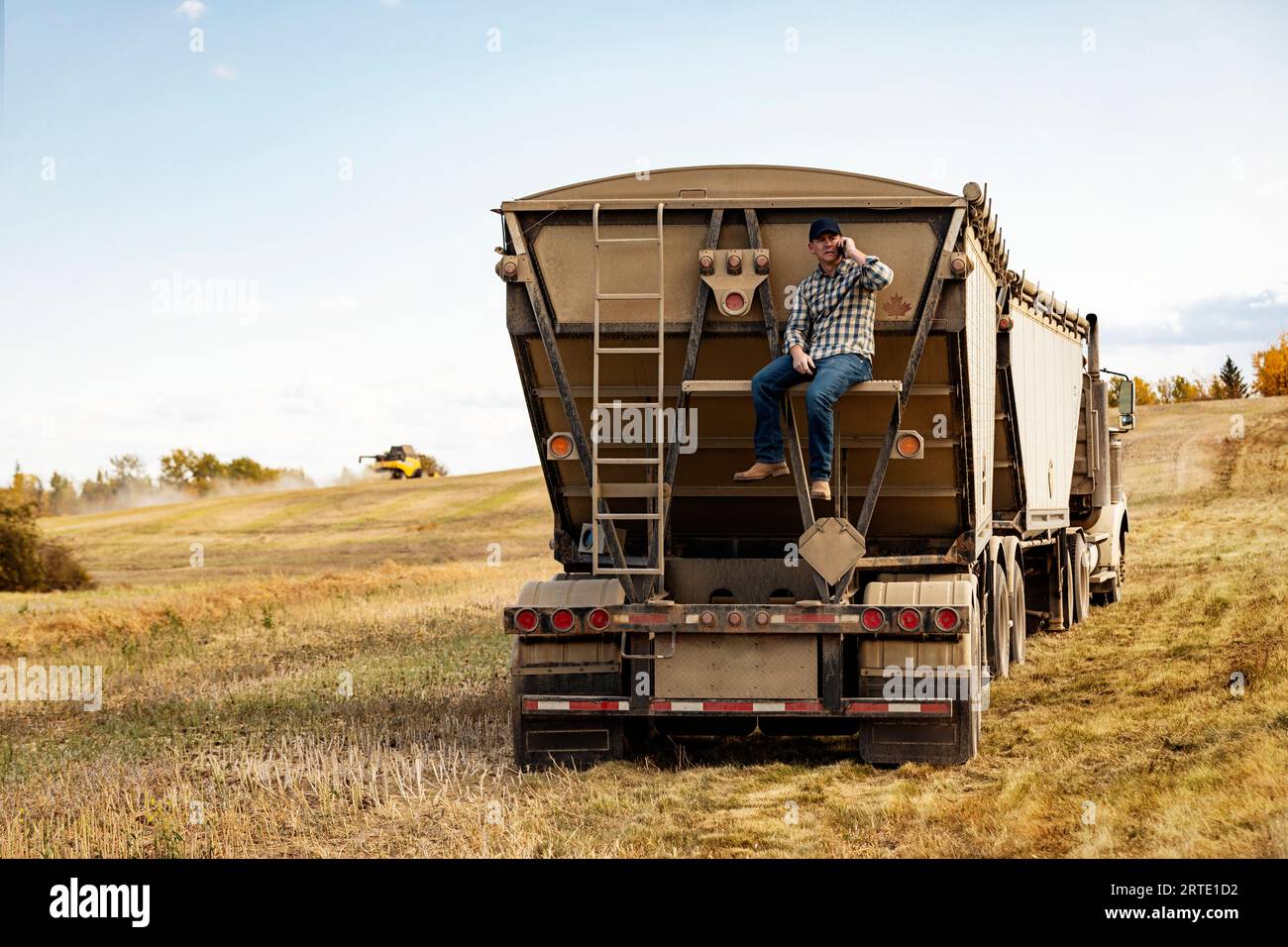 Ein Mann, der auf einem Smartphone spricht, während er hoch oben auf dem Rücken eines Getreidetransporters sitzt, seine Ladung Rapsöl überprüft und den Abschluss des... Stockfoto