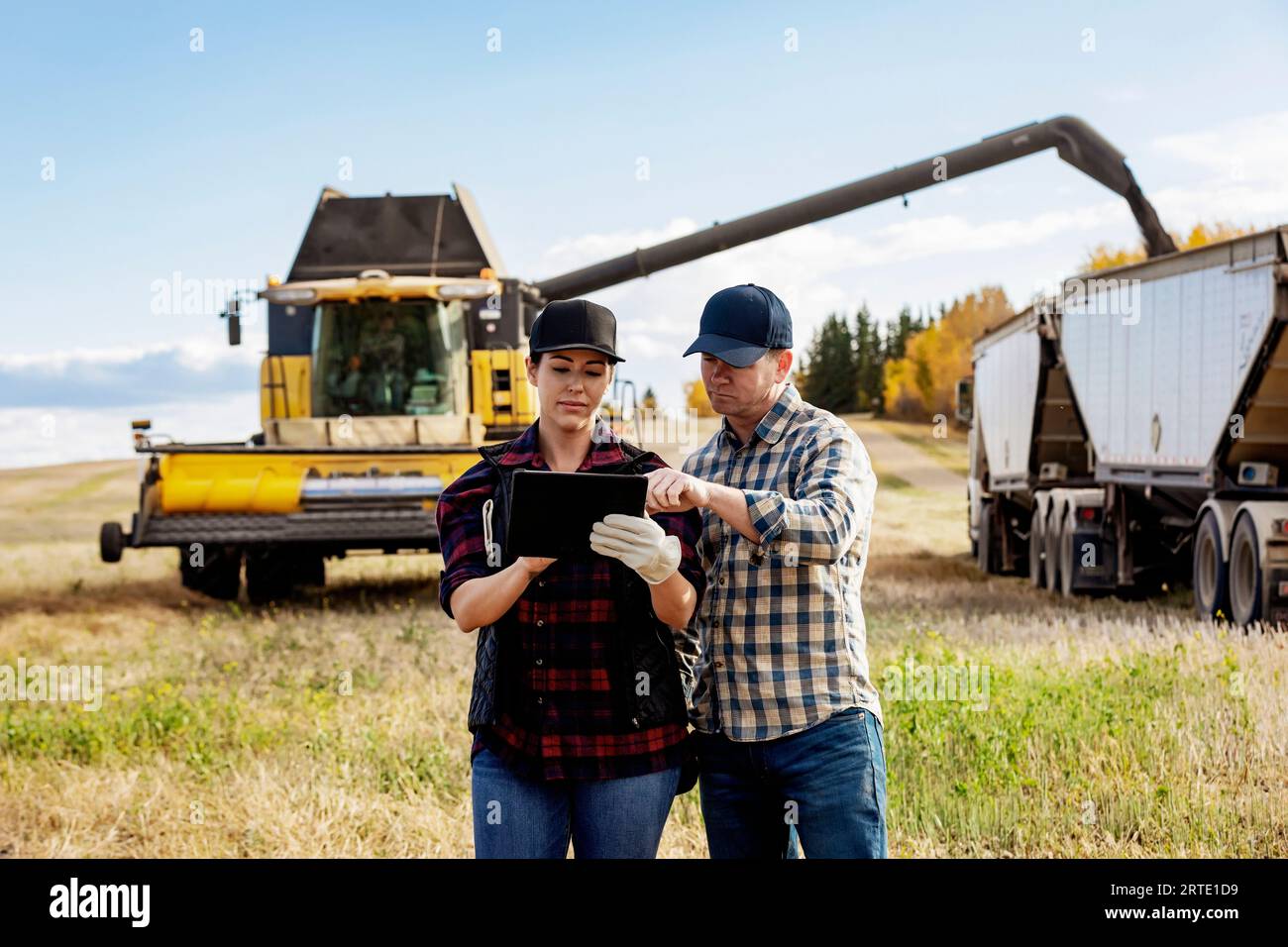 Nahaufnahme eines Mannes und einer Frau, die ein tragbares drahtloses Gerät zur Verwaltung und Überwachung ihrer Rapsernten verwenden, während der Mähdrescher Getreide in... Stockfoto